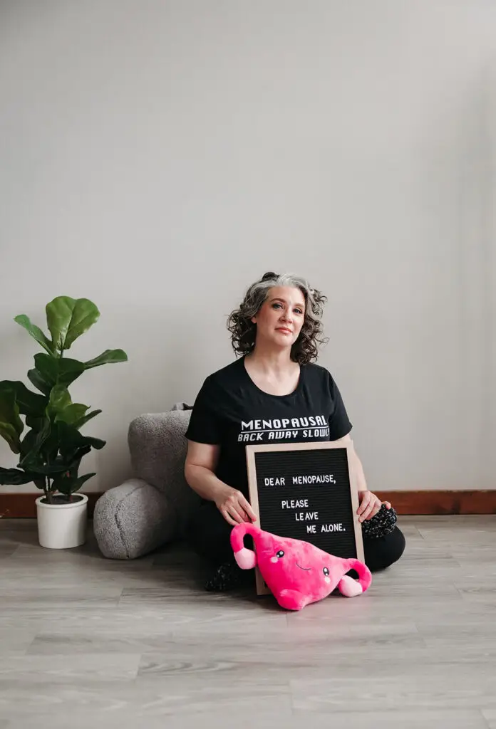 Coach Kristy sitting on the floor with a sign that says "Dear Menopause, please leave me alone" with a stuffed uterus in front of the sign and a plant in the background